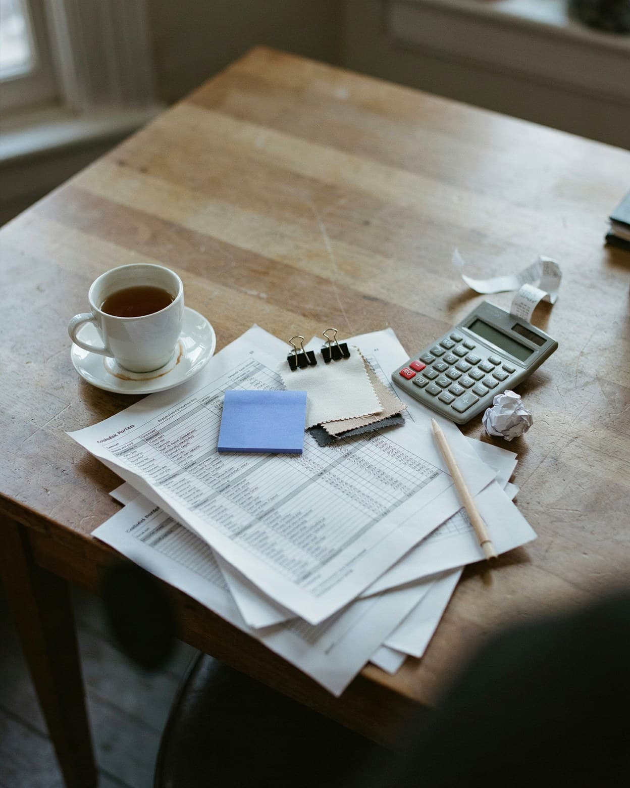 A costing desk at the end of the day: printed spreadsheets, a calculator, fabric swatches and one blue sticky note